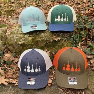 Pode incluir: Quatro bon&eacute;s de caminhoneiro em v&aacute;rias cores: verde-azulado, verde, azul marinho e azeitona. Cada bon&eacute; apresenta uma parte traseira em malha e um painel frontal bordado com uma linha de instrumentos musicais. Os bon&eacute;s exibem a marca "RICHARDSON" e o n&uacute;mero "115" no r&oacute;tulo.