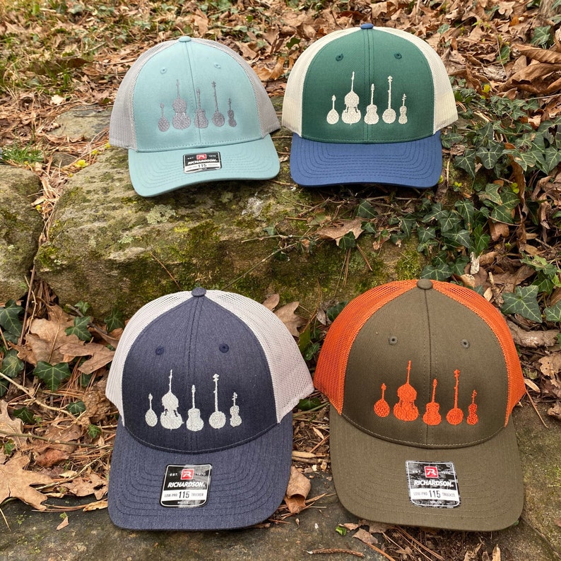 Pode incluir: Quatro bon&eacute;s de caminhoneiro em v&aacute;rias cores: verde-azulado, verde, azul marinho e azeitona. Cada bon&eacute; apresenta uma parte traseira em malha e um painel frontal bordado com uma linha de instrumentos musicais. Os bon&eacute;s exibem a marca "RICHARDSON" e o n&uacute;mero "115" no r&oacute;tulo.