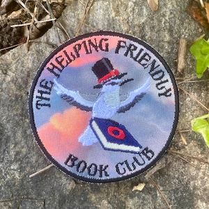 May include: Circular embroidered patch with the text "THE HELPING FRIENDLY BOOK CLUB." The patch features a blue bird wearing a top hat and holding a book. The background has a colorful sky design with shades of pink, orange, and blue.