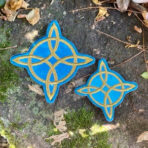 May include: Two embroidered patches with a Celtic knot design. The patches feature a light blue background with a gold-colored knot design. The patches are in the shape of a four-pointed star. One patch is larger than the other.