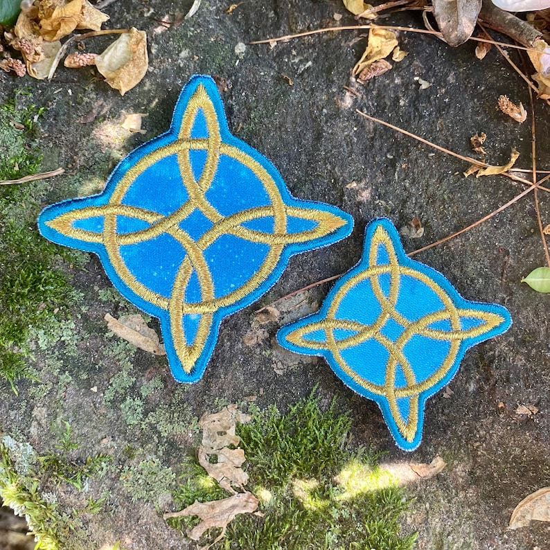 May include: Two embroidered patches with a Celtic knot design. The patches feature a light blue background with a gold-colored knot design. The patches are in the shape of a four-pointed star. One patch is larger than the other.