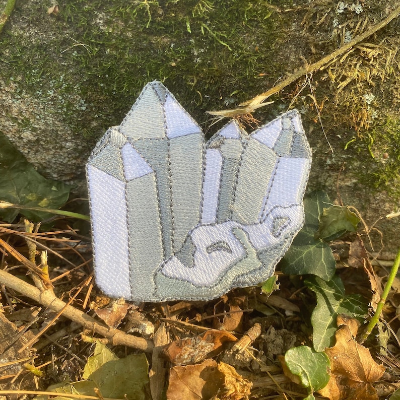 May include: Embroidered patch depicting a cluster of crystals in grey and white. The patch features a detailed design with geometric shapes, set against a natural background of leaves and moss.