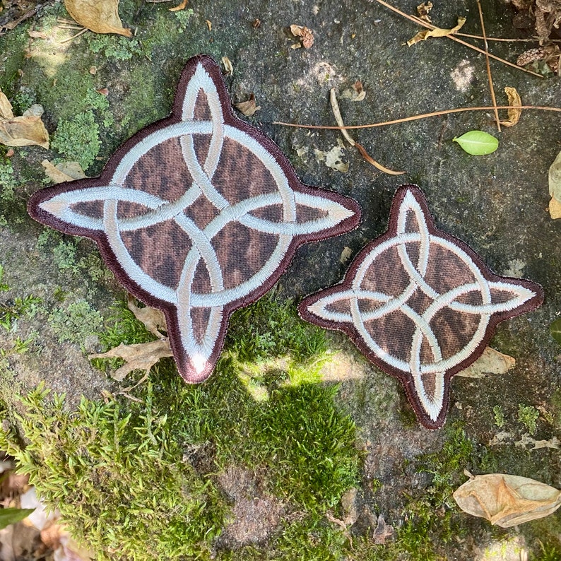 May include: Two brown and beige Celtic knot patches. The patches have a four-pointed star shape with a circular knot design in the center. The patches are embroidered with beige thread and have a brown background.