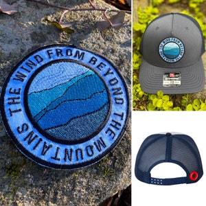 May include: A grey trucker cap with a circular embroidered patch depicting a mountain scene in shades of blue and the text "THE WIND FROM BEYOND THE MOUNTAINS." The cap has a black mesh back and a curved peak.