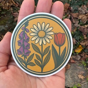 May include: A round sticker featuring a floral design. The sticker has a mustard yellow background with a white border. The design includes a purple foxglove, a white daisy, and a red tulip, with green leaves.