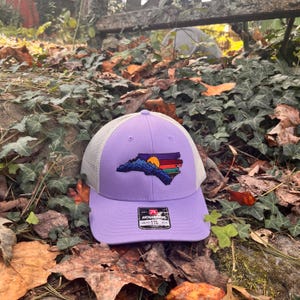 K&ouml;nnte beinhalten: Eine lila Baseballkappe mit wei&szlig;em Mesh-R&uuml;cken. Auf der Vorderseite befindet sich ein gesticktes Design des Umrisses des Bundesstaates North Carolina mit einer Bergkette, einem Sonnenuntergang und bunten Streifen.