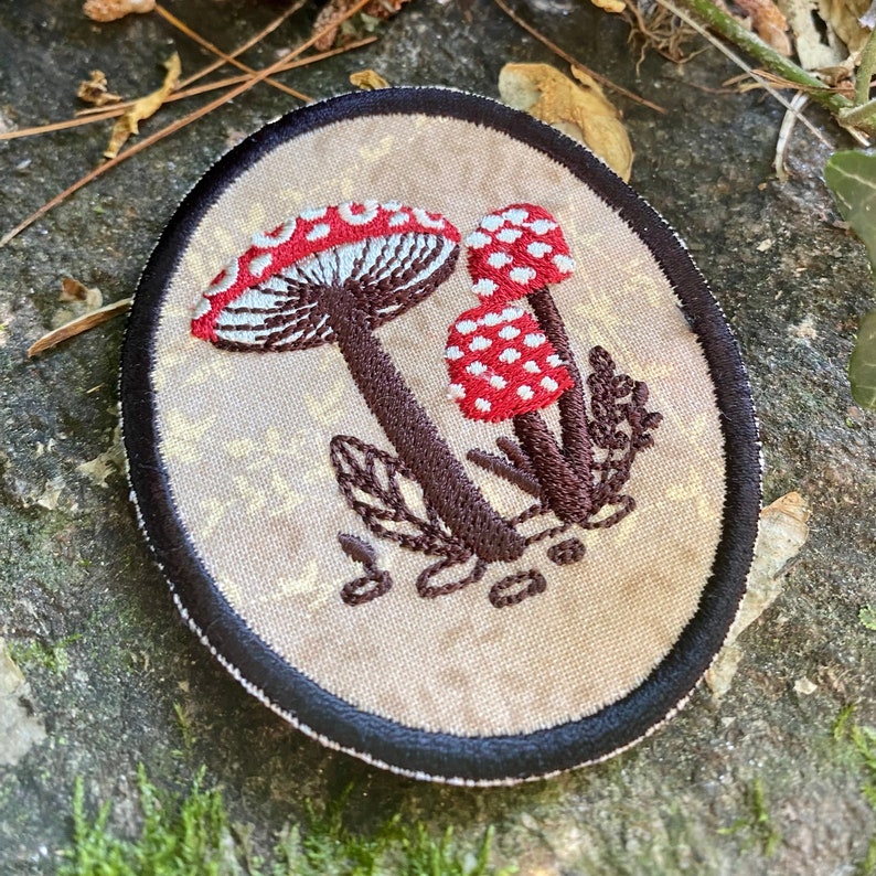 May include: Embroidered patch featuring three red and white spotted mushrooms with brown stems and leaves. The patch has a beige background with a black border. The patch is an oval shape.