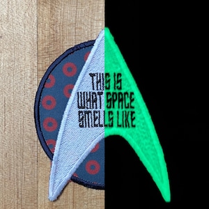 May include: Embroidered patch with a blue and red circular design and a light blue and green triangular shape. The text "THIS IS WHAT SPACE SMELLS LIKE" is embroidered on the patch. The green section glows in the dark.