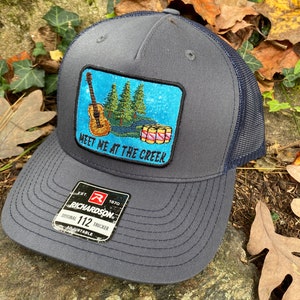 May include: Gray trucker hat with a navy blue mesh back and brim. The front patch features a guitar, trees, and the text "Meet Me At The Creek." The hat has an adjustable strap and a Richardson tag.