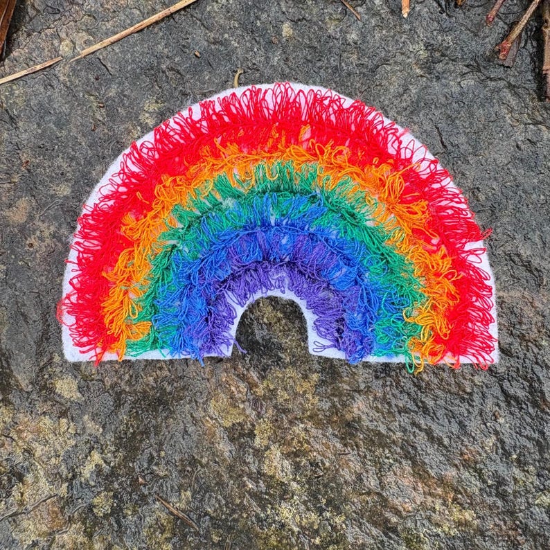 May include: A colorful, handmade rainbow patch with red, orange, yellow, green, blue, and purple yarn. The rainbow is stitched onto a white felt base. The patch is a cheerful, decorative item.