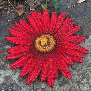 May include: A vibrant, red daisy-shaped textile with a yellow and brown center. The petals are a deep red with black outlines, creating a textured appearance. The daisy is placed on a gray, textured surface.