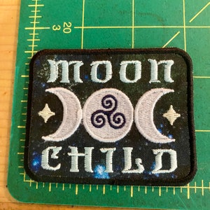 Op de afbeelding: Geborduurde patch met de woorden "MOON CHILD" in lichtblauw, met halve manen, een cirkelvormig symbool en sterren op een donkerblauwe achtergrond. De patch heeft een zwarte rand.