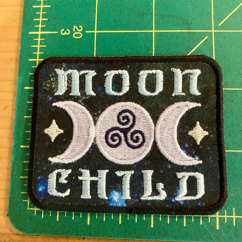 Op de afbeelding: Geborduurde patch met de woorden "MOON CHILD" in lichtblauw, met halve manen, een cirkelvormig symbool en sterren op een donkerblauwe achtergrond. De patch heeft een zwarte rand.