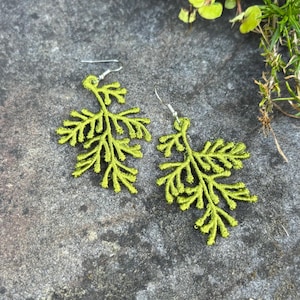 May include: A pair of lime green leaf-shaped earrings with silver-colored hooks. The earrings are made of a delicate, embroidered material, and are displayed on a textured gray surface. The earrings have a natural, botanical theme.