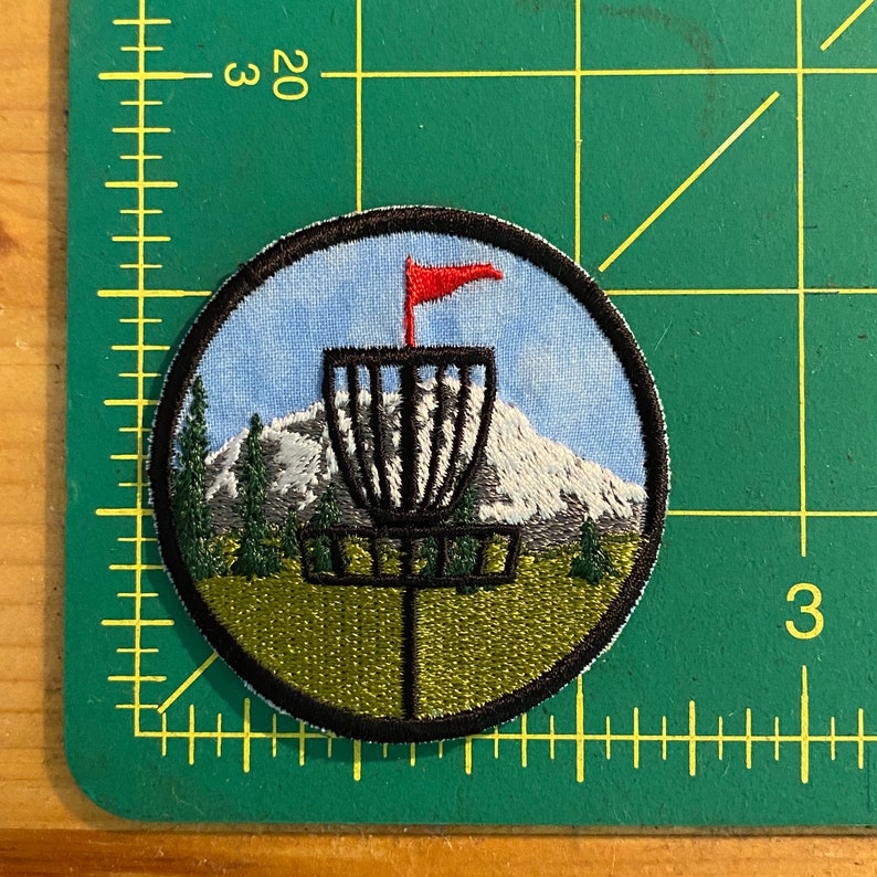 May include: Embroidered patch depicting a disc golf basket with a red flag, set against a mountain range and evergreen trees. The circular patch has a black border and is approximately 7.6 cm in diameter.