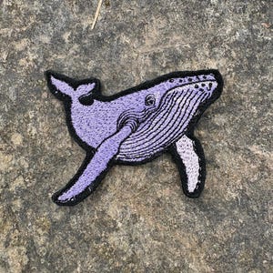 May include: Embroidered patch featuring a purple whale with black outline. The whale has detailed stitching and a white underside. The patch is designed to be ironed on or sewn onto fabric. Ideal for adding a decorative touch to clothing or accessories.