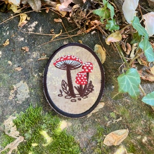 May include: An oval patch featuring an embroidered design of three red and white spotted mushrooms with brown stems and leaves. The patch has a tan background with a gold floral pattern and a black border. The patch is likely for clothing or accessories.