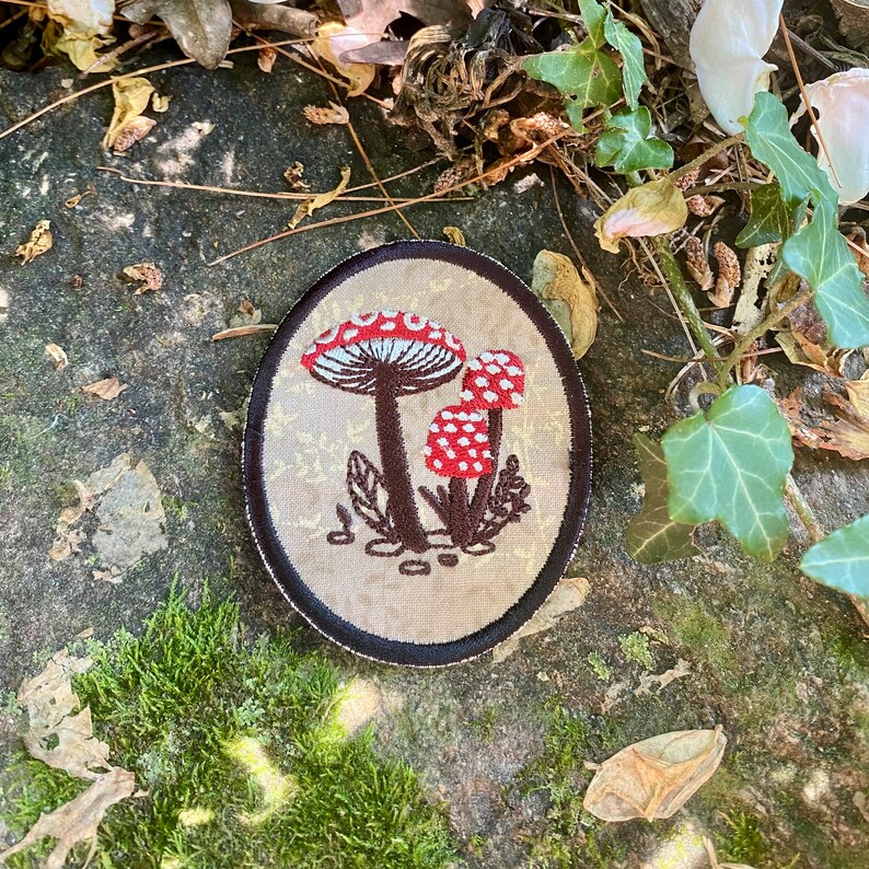 May include: An oval patch featuring an embroidered design of three red and white spotted mushrooms with brown stems and leaves. The patch has a tan background with a gold floral pattern and a black border. The patch is likely for clothing or accessories.