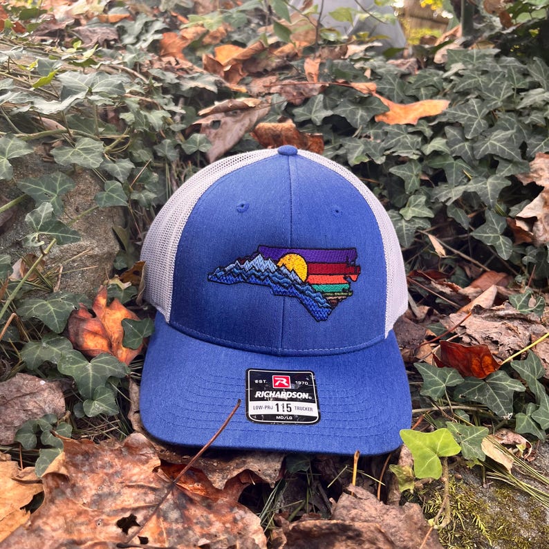 K&ouml;nnte beinhalten: Blaue Trucker-Cap mit wei&szlig;em Mesh-R&uuml;cken. Auf der Vorderseite befindet sich ein gesticktes Design des Umrisses des Bundesstaates North Carolina mit einer Bergkette, einem Sonnenuntergang und bunten Streifen. Die Kappe hat einen gebogenen Schirm.
