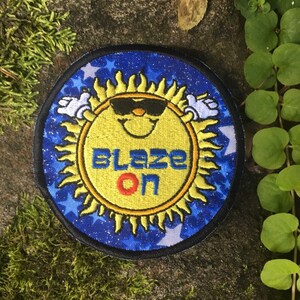 Puede incluir: Parche bordado con un sol sonriente con gafas de sol y el texto "BLAZE ON". El sol es amarillo con un contorno negro, sobre un fondo azul con estrellas blancas. El parche tiene un borde negro.