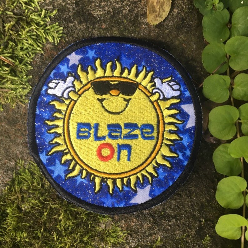 Puede incluir: Parche bordado con un sol sonriente con gafas de sol y el texto "BLAZE ON". El sol es amarillo con un contorno negro, sobre un fondo azul con estrellas blancas. El parche tiene un borde negro.