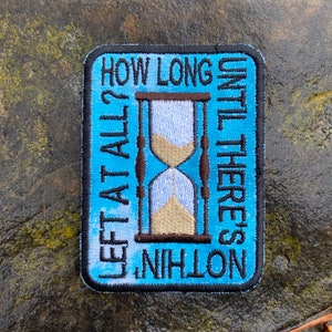 May include: Embroidered patch with a light blue background and black border. The patch features an hourglass design with the text "HOW LONG LEFT AT ALL? UNTIL THERE'S NOTHING". The hourglass has tan sand.