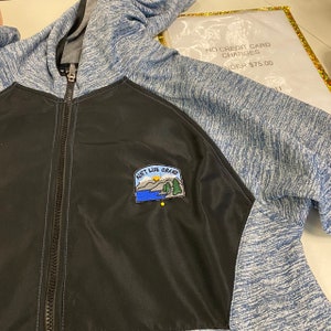 May include: A black and blue zip-up hooded jacket with a landscape patch that reads "Aunt Life Grand". The jacket has a black front panel and blue sleeves and hood. The zipper is silver.