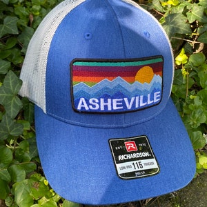 Peut inclure: Casquette de camionneur bleue avec un dos en filet blanc. L'avant pr&eacute;sente un &eacute;cusson avec une sc&egrave;ne de montagne et le mot "Asheville". La casquette a une visi&egrave;re incurv&eacute;e et une &eacute;tiquette Richardson 115 Low-Pro Trucker.