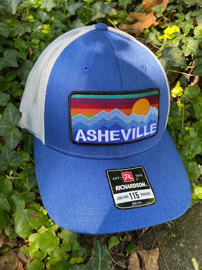 Peut inclure: Casquette de camionneur bleue avec un dos en filet blanc. L'avant pr&eacute;sente un &eacute;cusson avec une sc&egrave;ne de montagne et le mot "Asheville". La casquette a une visi&egrave;re incurv&eacute;e et une &eacute;tiquette Richardson 115 Low-Pro Trucker.