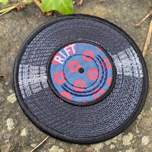 May include: Embroidered patch in the shape of a vinyl record. The record is primarily dark blue with a black border. The center label is blue with red donut-shaped designs and the word "RIFT".
