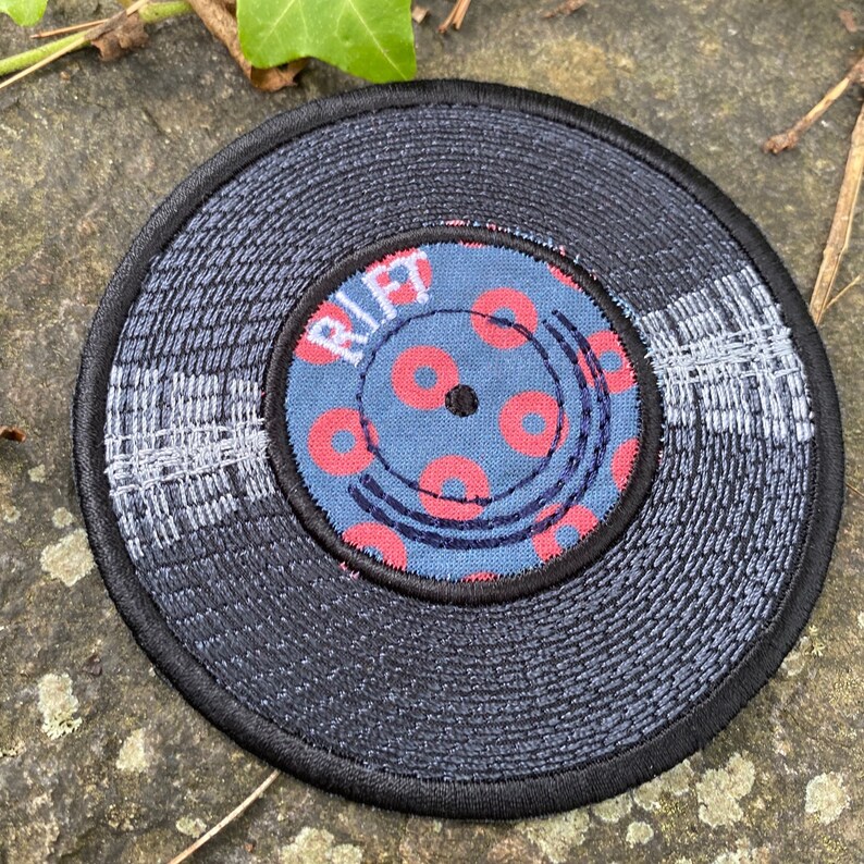 May include: Embroidered patch in the shape of a vinyl record. The record is primarily dark blue with a black border. The center label is blue with red donut-shaped designs and the word "RIFT".