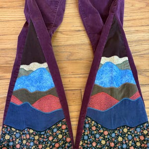 Op de afbeelding: Pruimkleurige corduroy broek met uitlopende pijpen. De uitlopende secties hebben een berglandschapsontwerp in tinten blauw, bruin, rood en bloempatronen. De broek ligt op een houten oppervlak.