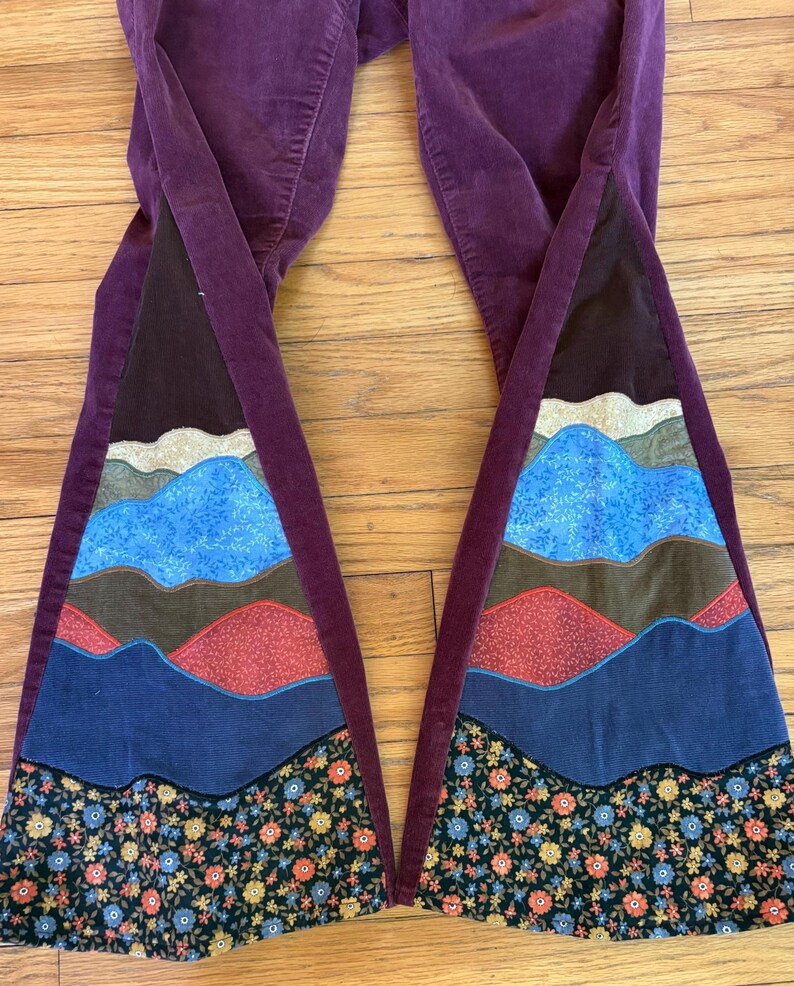 Op de afbeelding: Pruimkleurige corduroy broek met uitlopende pijpen. De uitlopende secties hebben een berglandschapsontwerp in tinten blauw, bruin, rood en bloempatronen. De broek ligt op een houten oppervlak.