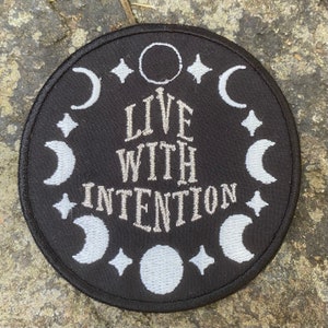 May include: Circular black patch with white embroidered design. The patch features the text "LIVE WITH INTENTION" surrounded by crescent moons, stars, and a circle. Suitable for clothing or accessories.
