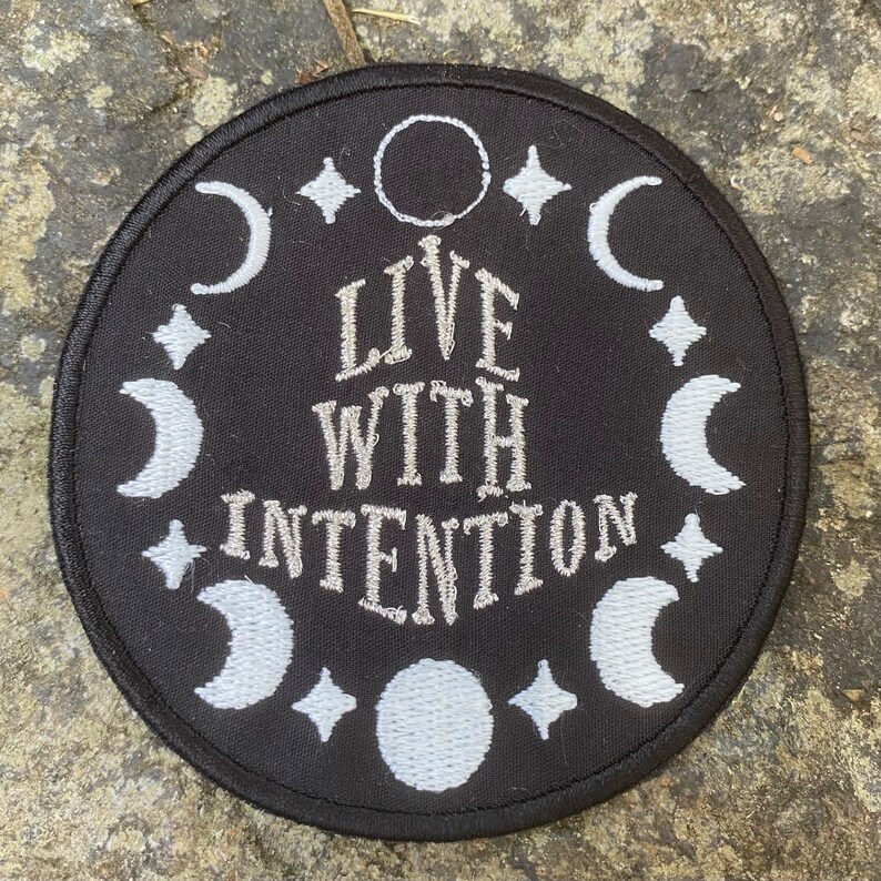 May include: Circular black patch with white embroidered design. The patch features the text "LIVE WITH INTENTION" surrounded by crescent moons, stars, and a circle. Suitable for clothing or accessories.