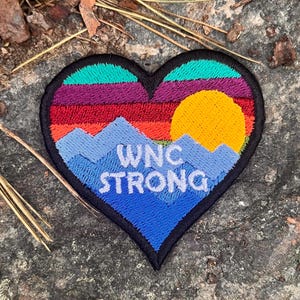 May include: Heart-shaped embroidered patch with a mountain and sunset design. The patch features the text "WNC STRONG" in white, with a blue mountain range, a yellow sun, and a colorful sunset. The patch is outlined in black.