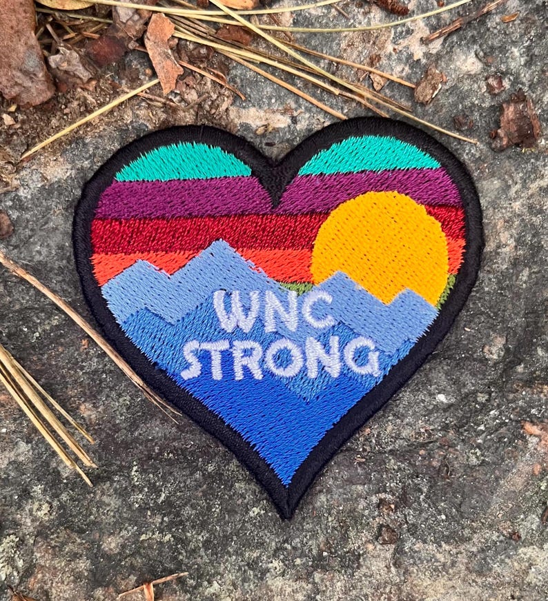 以下が含まれることがあります： 山と夕日のデザインが施された、ハート型の刺繍パッチ。「WNC STRONG」の文字が白で、青い山脈、黄色の太陽、カラフルな夕日が特徴です。パッチは黒で縁取られています。