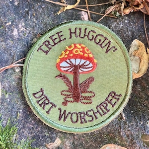 May include: A round, olive green embroidered patch with the words "TREE HUGGIN'" and "DIRT WORSHIPER" in brown thread. A colorful mushroom with a red cap, yellow spots, and white gills is in the center.