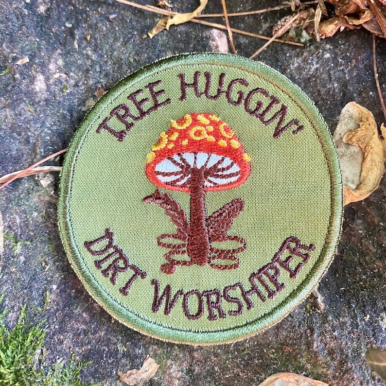 Pu&ograve; includere: Una toppa rotonda ricamata verde oliva con le parole "TREE HUGGIN'" e "DIRT WORSHIPER" in filo marrone. Un fungo colorato con cappello rosso, macchie gialle e lamelle bianche &egrave; al centro.