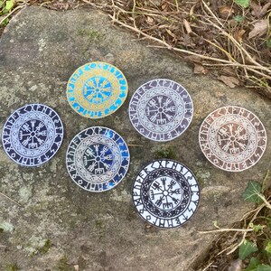 May include: Seven circular coasters featuring a Viking compass design. The coasters display a range of colours, including blue, gold, brown, and black, with white detailing. They are arranged on a textured, grey stone surface.