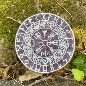 May include: A circular patch with a dark purple background and white embroidered Viking symbol. The symbol is surrounded by runes and intricate knotwork. The patch is placed on a bed of moss and leaves.