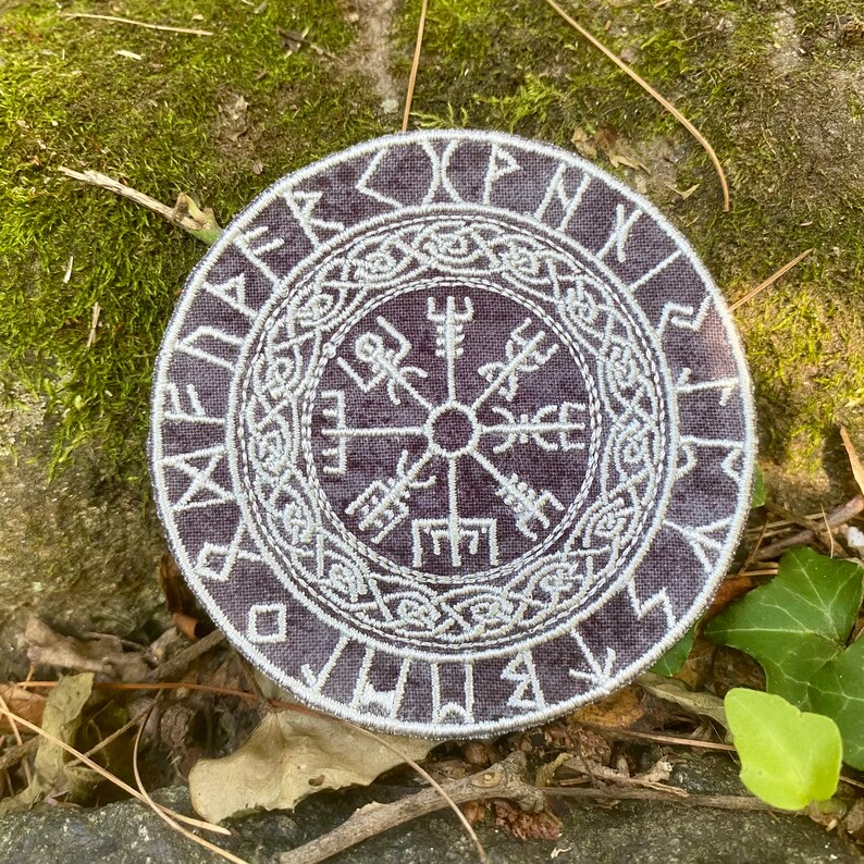 May include: A circular patch with a dark purple background and white embroidered Viking symbol. The symbol is surrounded by runes and intricate knotwork. The patch is placed on a bed of moss and leaves.