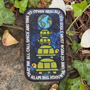 May include: Rectangular patch with a dark blue background, featuring a stack of four green turtles supporting a globe. The patch has white text that reads "THAT WE ALL CALL SPACE AND TIME" and "OUR SOULS MUST ROAM TO AND THROUGH THE MYTH."