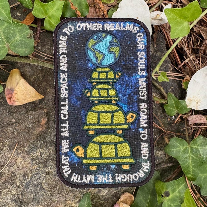 May include: Rectangular patch with a dark blue background, featuring a stack of four green turtles supporting a globe. The patch has white text that reads "THAT WE ALL CALL SPACE AND TIME" and "OUR SOULS MUST ROAM TO AND THROUGH THE MYTH."