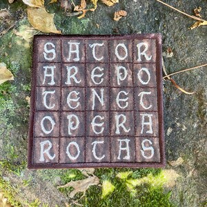 May include: A square, brown embroidered patch with the text "SATOR AREPO TENET OPERA ROTAS" in white, stylized lettering. The patch is framed with a darker brown border. The background is a natural setting with stone, moss, and fallen leaves.
