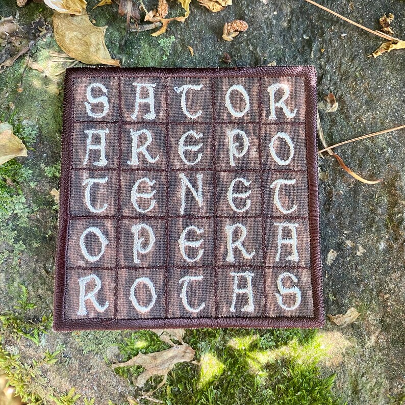 May include: A square, brown embroidered patch with the text "SATOR AREPO TENET OPERA ROTAS" in white, stylized lettering. The patch is framed with a darker brown border. The background is a natural setting with stone, moss, and fallen leaves.