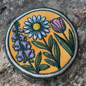 Pu&ograve; includere: Toppa ricamata con un design floreale, tra cui una margherita, un tulipano e fiori di digitale. La toppa ha uno sfondo giallo con dettagli verdi e rosa e un bordo cucito. La toppa &egrave; di forma rotonda.
