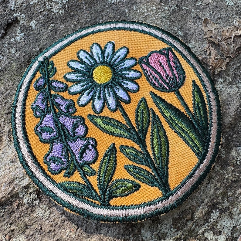 Pu&ograve; includere: Toppa ricamata con un design floreale, tra cui una margherita, un tulipano e fiori di digitale. La toppa ha uno sfondo giallo con dettagli verdi e rosa e un bordo cucito. La toppa &egrave; di forma rotonda.