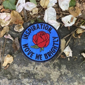 May include: A round, blue embroidered patch with the words "INSPIRATION, MOVE ME BRIGHTLY" encircling a red rose. The patch is on a gray stone surface, surrounded by dried leaves and flower petals.