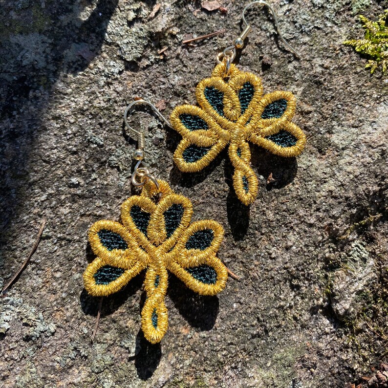 Pode incluir: Um par de brincos de trevo bordados em dourado com detalhes em preto. Os brincos s&atilde;o feitos com fio met&aacute;lico e t&ecirc;m fecho de gancho. Os brincos s&atilde;o exibidos em uma superf&iacute;cie cinza texturizada.